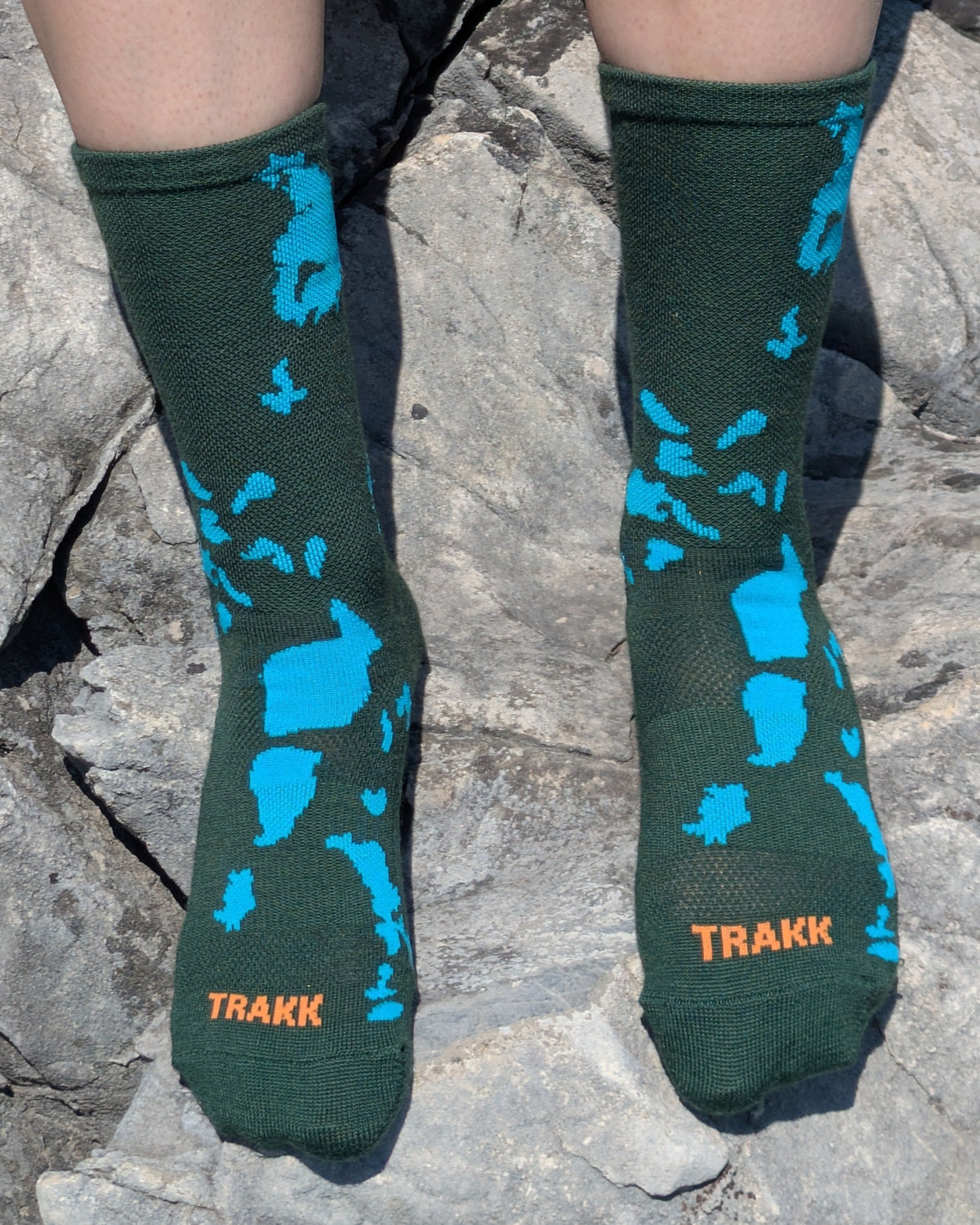 A person sitting on gray rocks wearing dark green socks with bright blue abstract shapes and orange TRAKK branding near the toes.